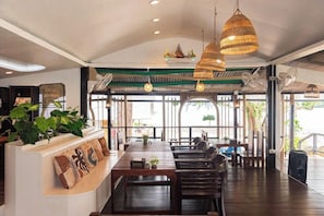 An indoor dining area with a long wooden table, multiple chairs, hanging wicker lamps, and a view through large windows.