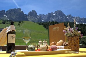 A table set with a bottle of wine, a glass, a menu, and various cheeses and bread, with a mountain view in the background. - Monte Croce Hotel Kreuzberg (Sesto)