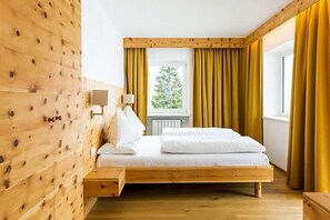 A wooden bed with a headboard, a window with yellow curtains, and a view of greenery outside. - Monte Croce Hotel Kreuzberg (Sesto)
