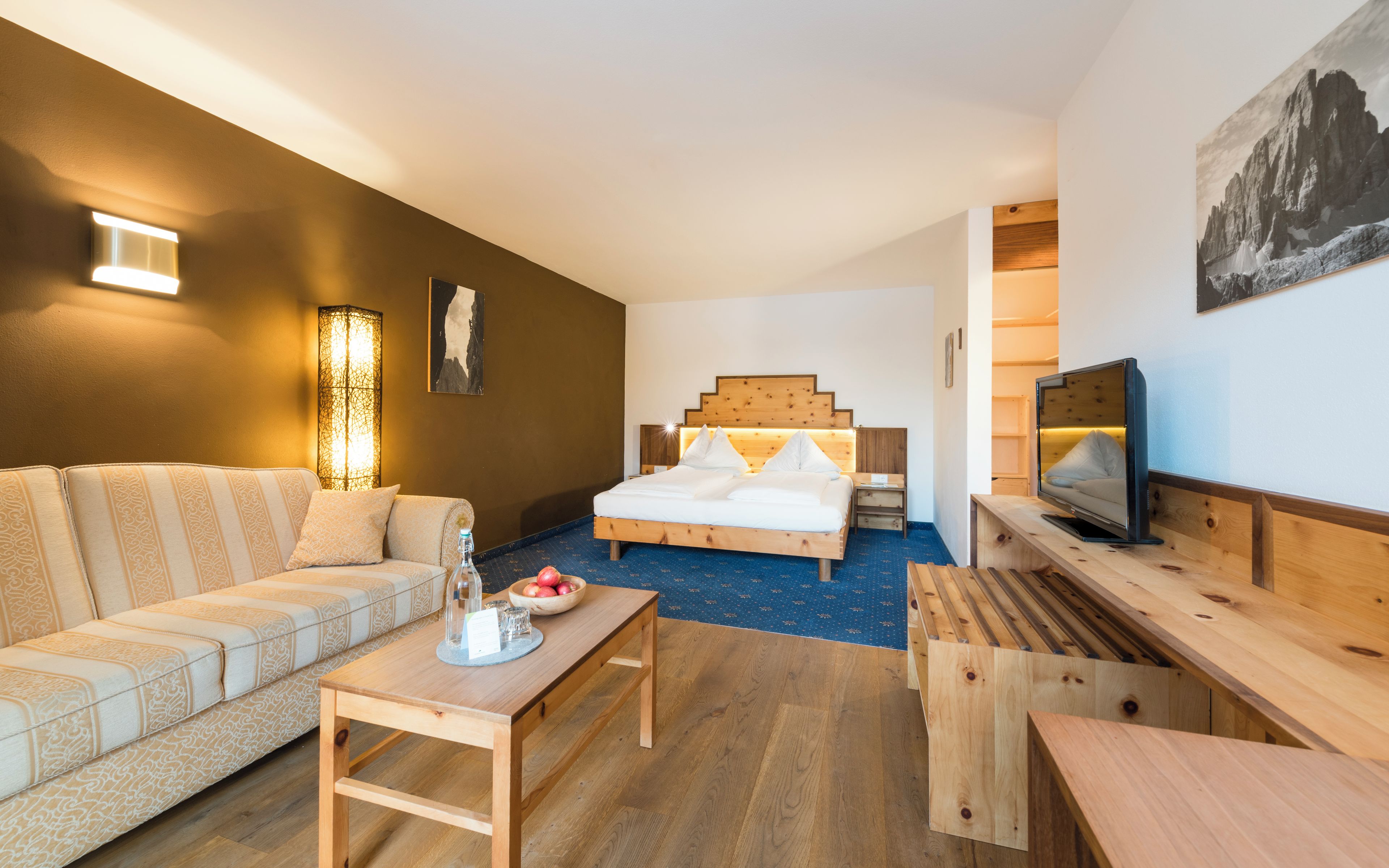 A modern hotel room with a wooden bed, a sofa, a TV, and a dining table.