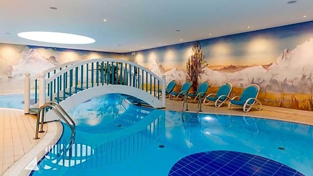 Una piscina interior con una escalera curva, sillones de descanso y un mural de un paisaje montañoso.