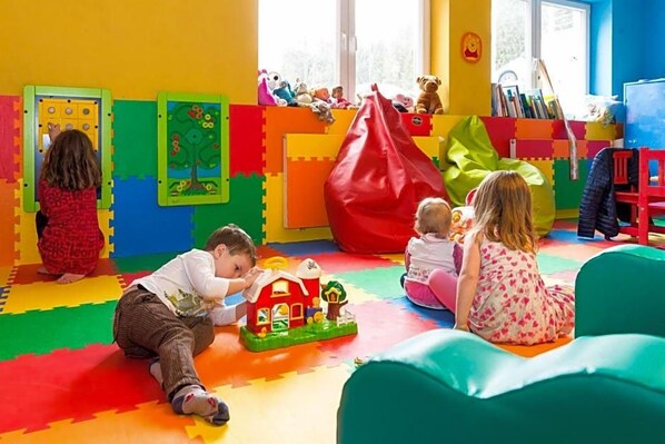 Een vrolijk speelgebied met foammatten, een speelhuisje en spelende kinderen.
