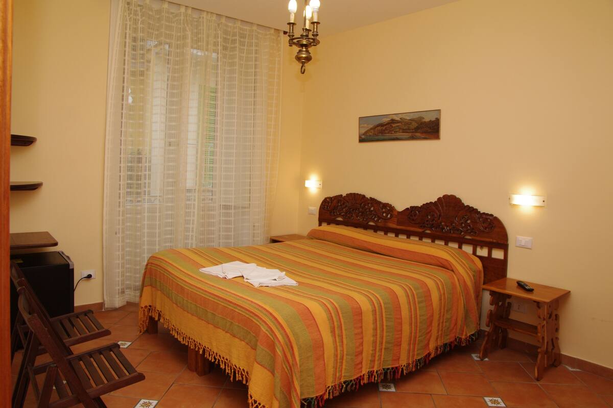 A bedroom with a bed, a wooden nightstand, a chair, a desk, and a window with curtains.