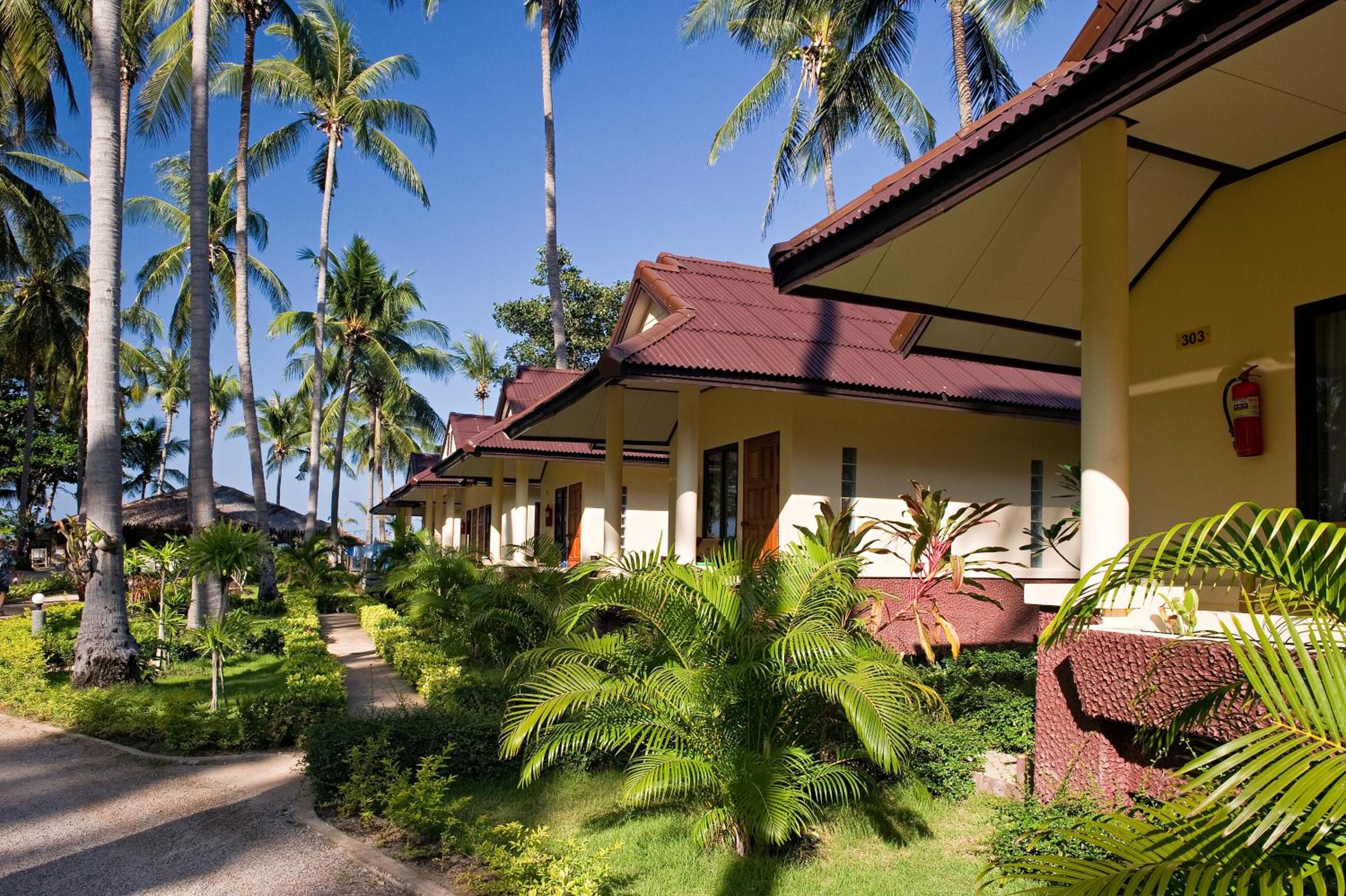A row of bungalow-style buildings with a thatched roof, surrounded by lush tropical vegetation and palm trees.