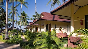 A row of bungalow-style buildings with a thatched roof, surrounded by lush tropical vegetation and palm trees.