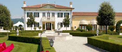 A large, white building with a central entrance, surrounded by a well-manicured garden with hedges and a fountain.
