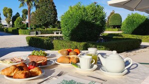 Un coin petit-déjeuner en plein air, avec des croissants, du pain, des fruits et du thé, disposés sur une table dans un jardin où les haies et les arbres sont soigneusement taillés.