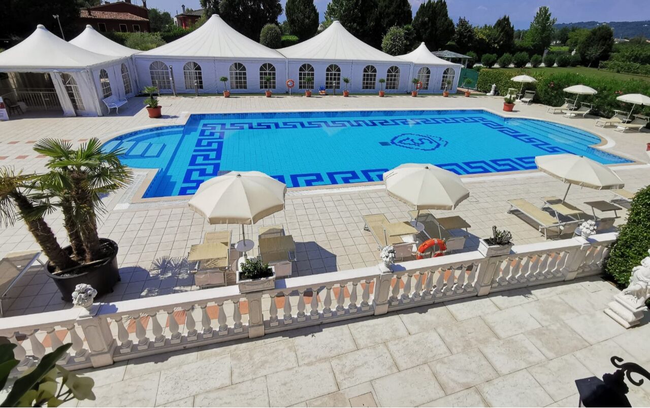 A swimming pool with a geometric pattern, surrounded by lounge chairs and umbrellas, with white tents in the background.