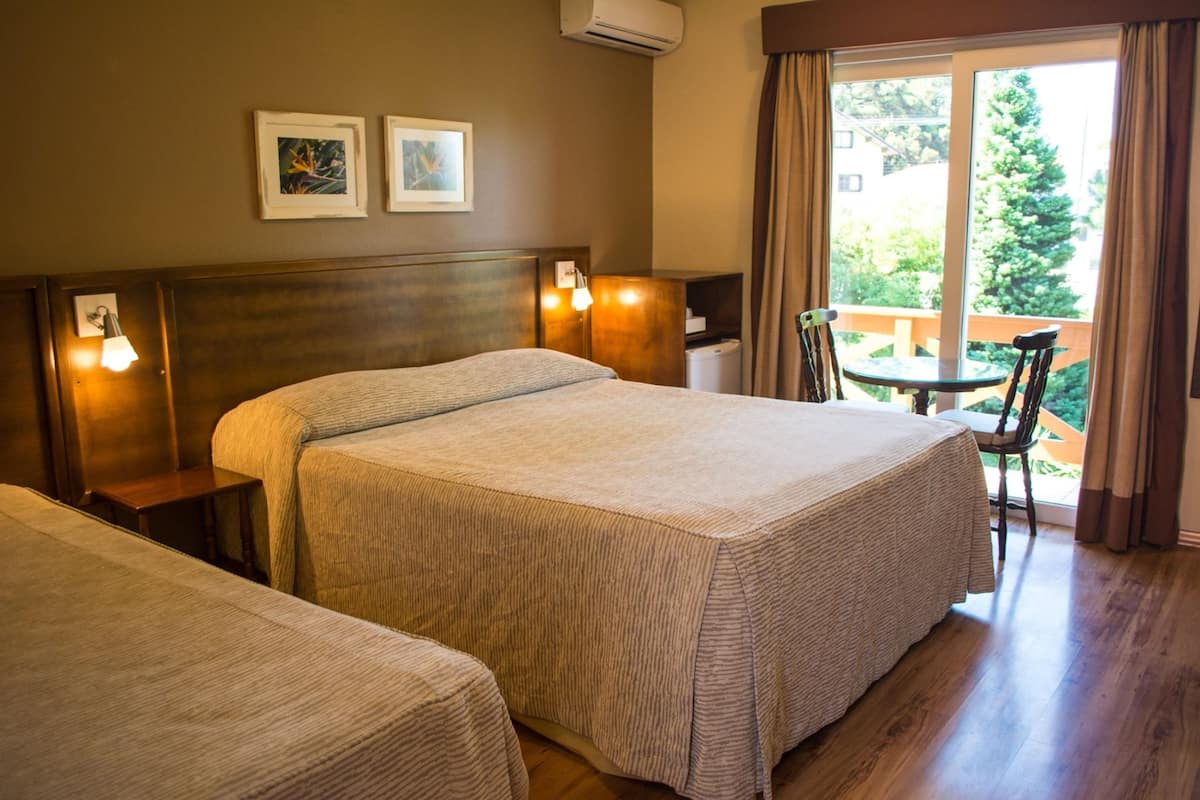 Habitación de hotel con dos camas, una mesita, una silla y un balcón con vistas al follaje.