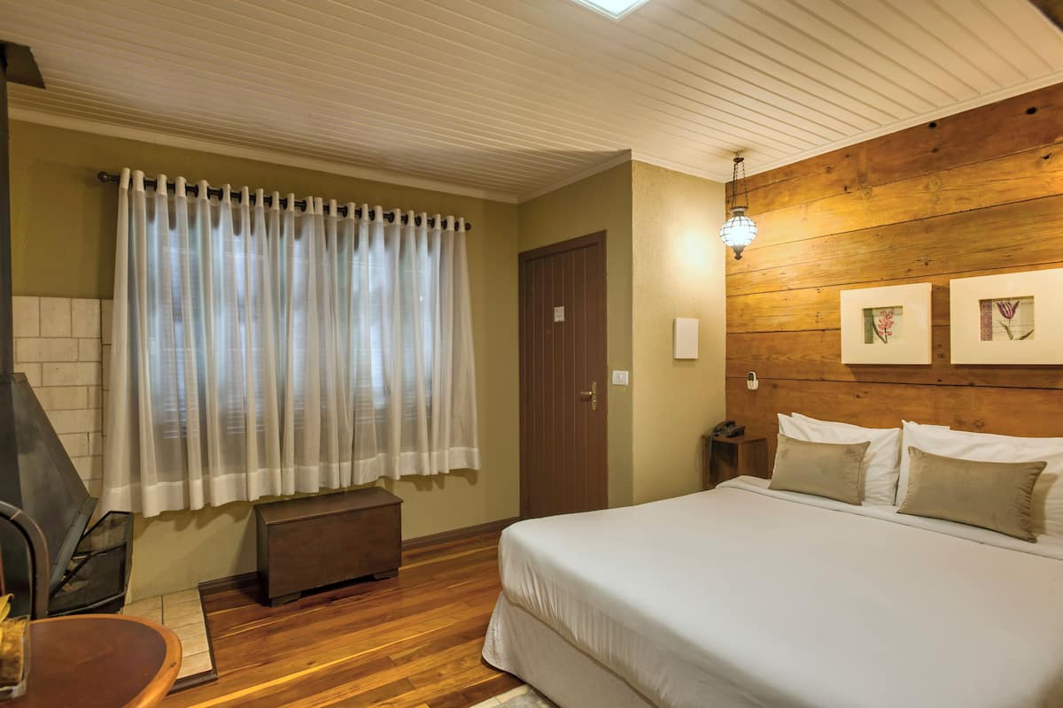 Un dormitorio con cabecera de madera, una cama grande con ropa de cama blanca, un banco de madera, una chimenea y una ventana con cortinas de gasa.