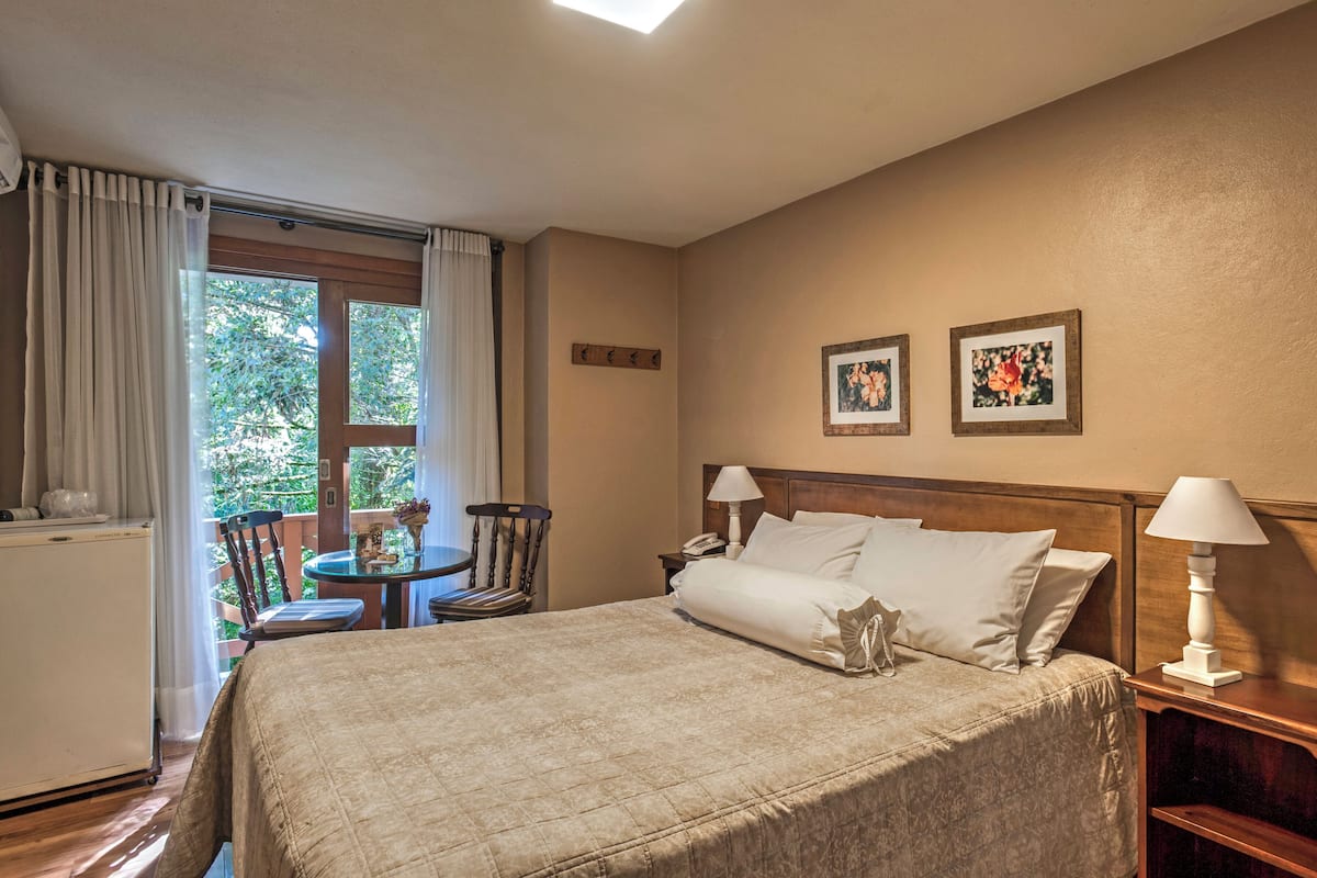 Habitación de hotel con cama, mesitas de noche, una silla, una pequeña mesa con un florero y una ventana con cortinas.