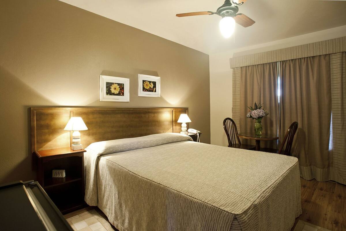 Habitación de hotel con una cama, dos lámparas de noche, un ventilador de techo, una mesita con un florero y dos cuadros en la pared.