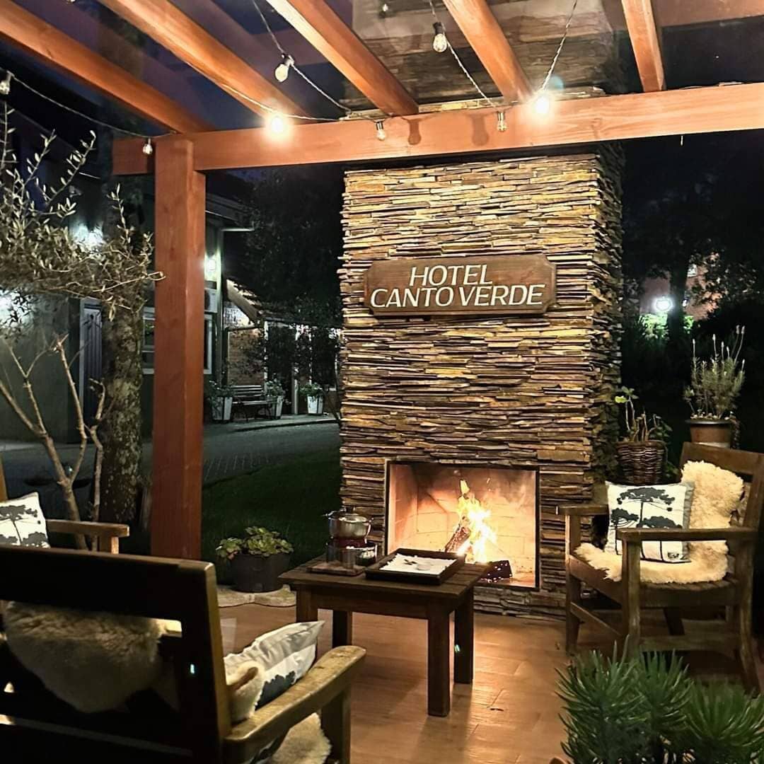 A cozy outdoor seating area with a stone fireplace labeled 'HOTEL CANTO VERDE,' surrounded by wooden furniture and potted plants.
