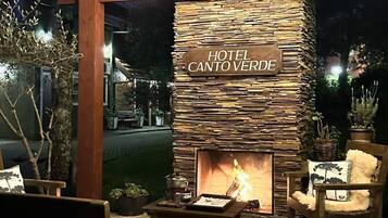 A cozy outdoor seating area with a stone fireplace labeled 'HOTEL CANTO VERDE,' surrounded by wooden furniture and potted plants.