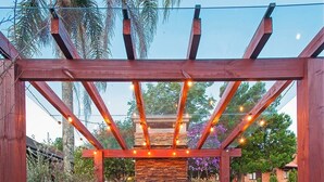 Una pergola in legno con un camino in pietra e luci a tendina, circondata da un giardino con una panchina e piante in vaso.