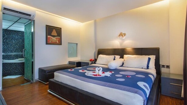 A modern hotel room with a large bed, a bedside table, a nightstand, and a bathroom with a shower and a tiled wall.