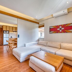 A modern living room with a sectional sofa, a wooden coffee table, and a kitchen area with a microwave and refrigerator.
