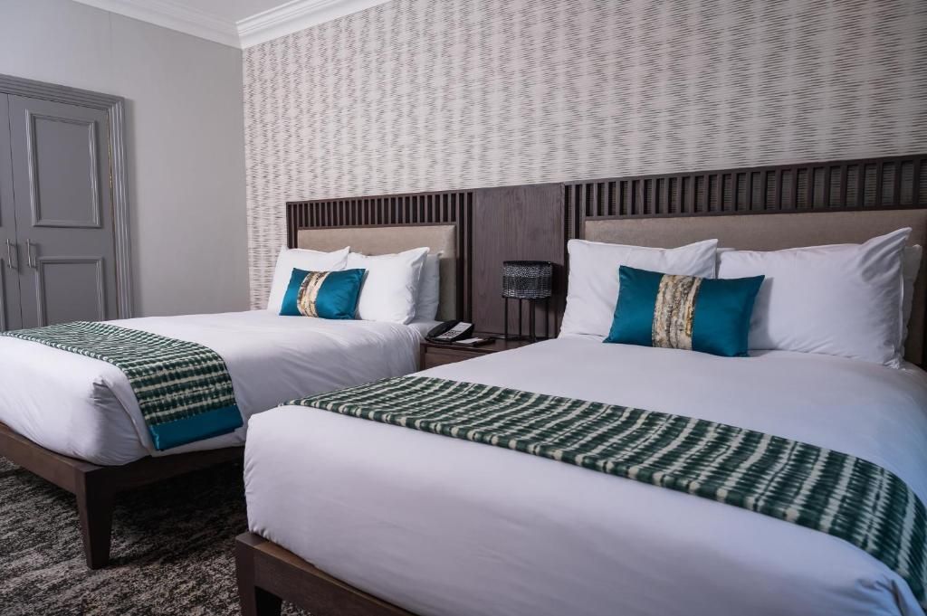 A hotel room with two beds, each with white bedding and green throw blankets. There is a wooden headboard, a bedside table with a lamp, and a telephone.