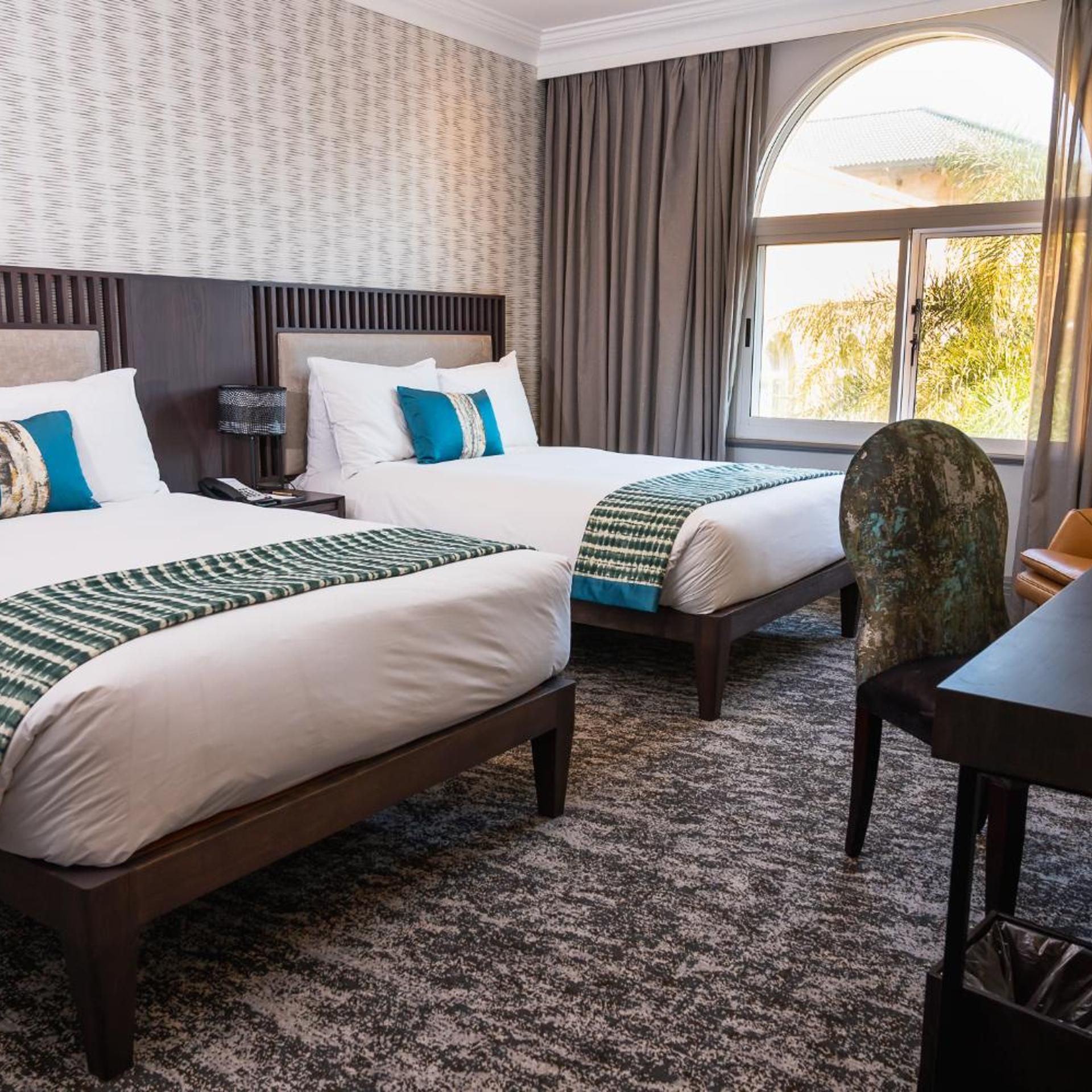 Une chambre d’hôtel avec deux lits, un bureau, une chaise et une grande fenêtre avec des rideaux.
