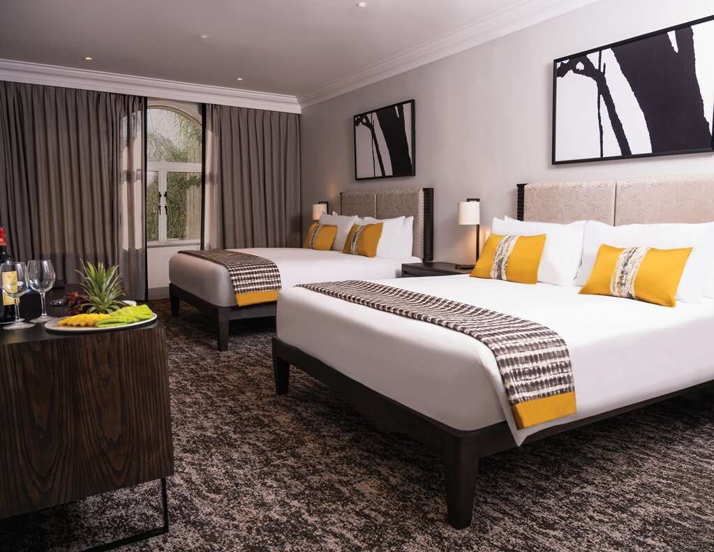 A hotel room with two beds, a wooden side table, and a large window with curtains.