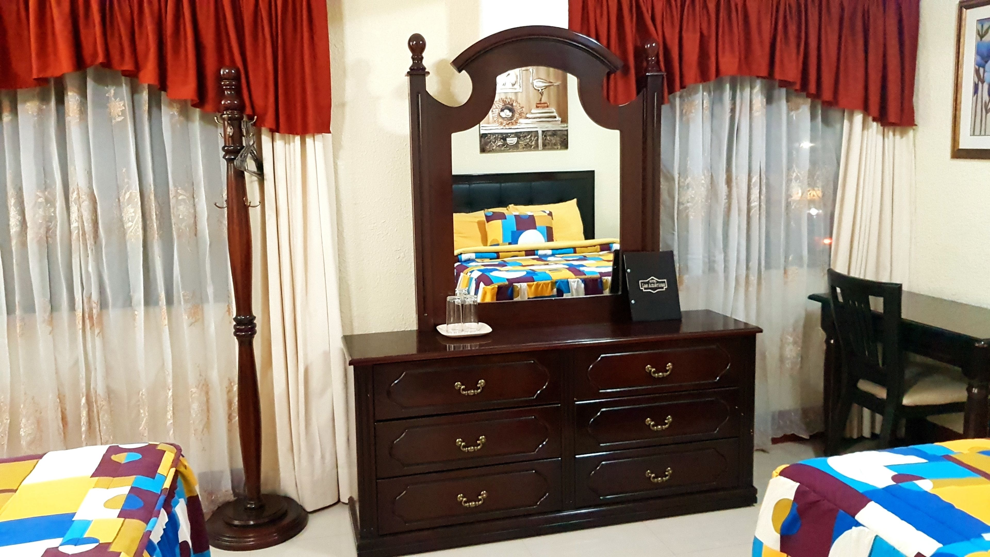 A bedroom with a wooden dresser, a mirror, a bed with colorful bedding, and a desk with a chair.