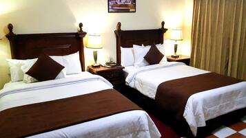 A hotel room with two beds, each with a wooden headboard, white bedding, and a brown throw blanket. There are bedside tables with lamps and a clock.