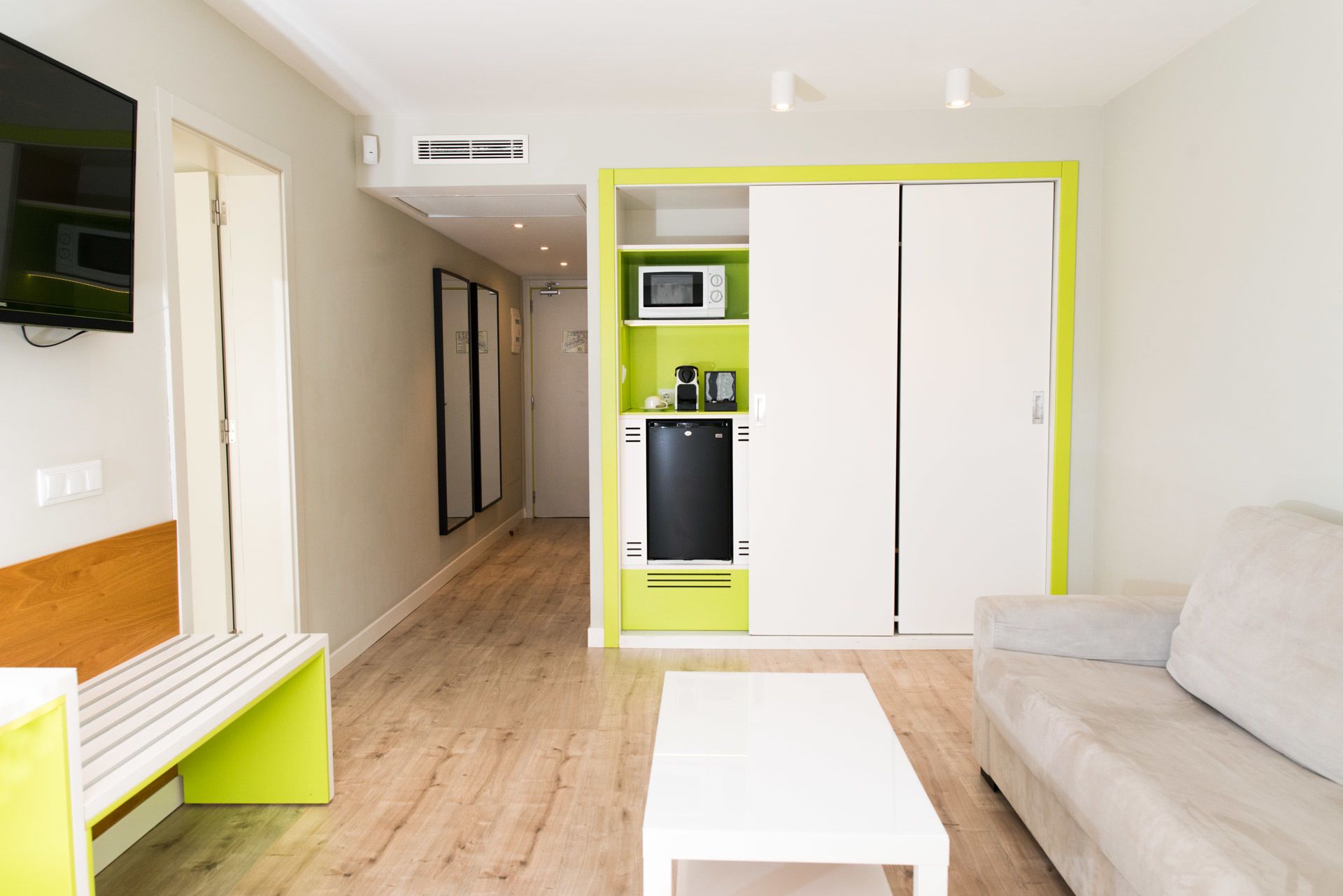 A compact living space with a kitchenette, a flat-screen TV mounted on the wall, and a sofa.