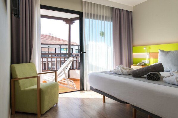 Habitación de hotel con cama, una silla, balcón con barandilla y vistas a edificios.
