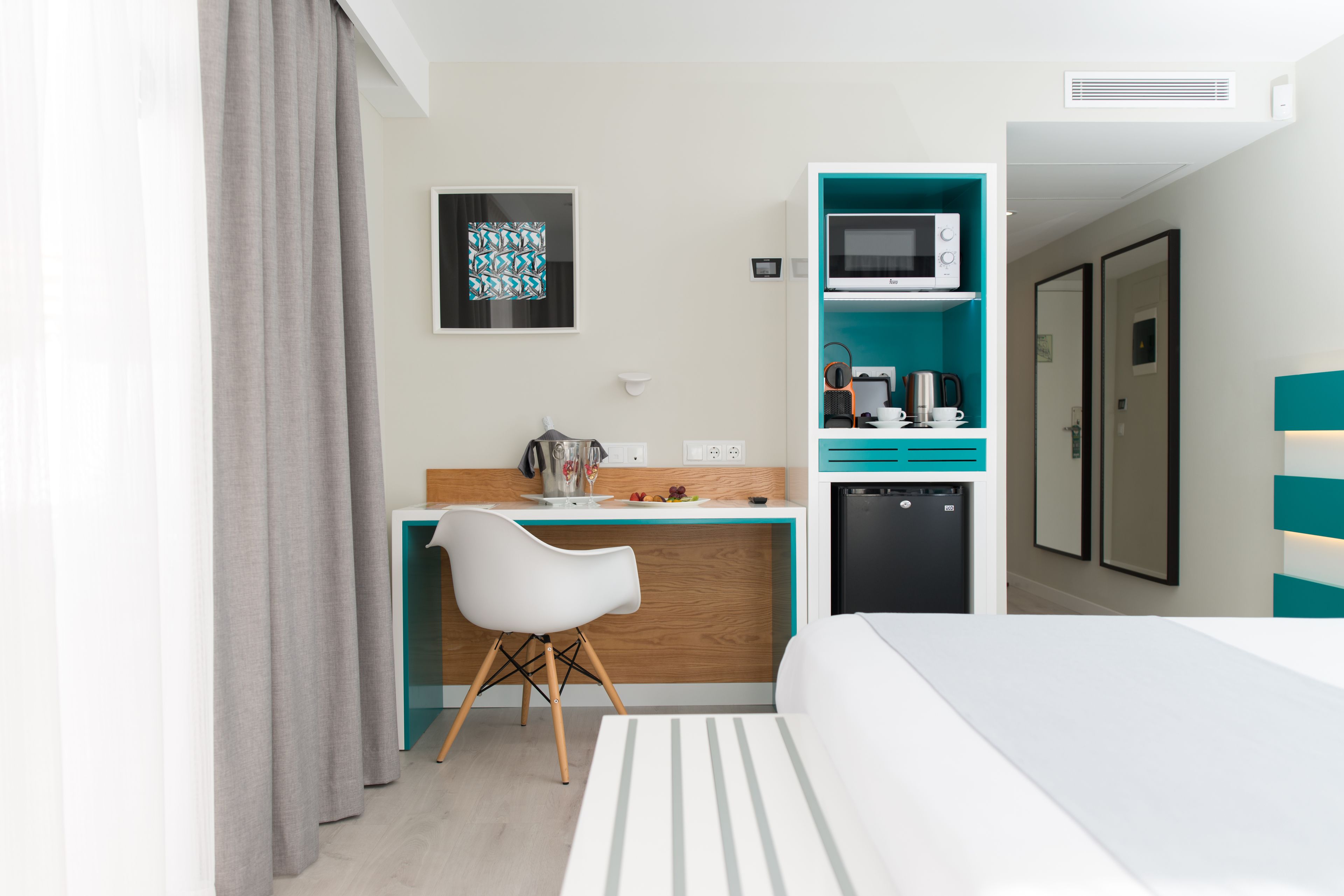Une chambre d’hôtel moderne équipée d’un lit, d’un bureau avec une chaise, d’un four à micro-ondes, d’une machine à café et d’un petit réfrigérateur.