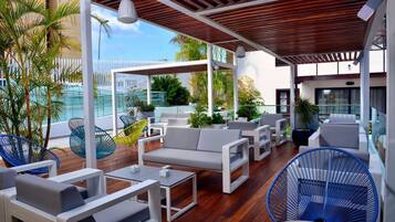 Un área de estar techada al aire libre con mobiliario moderno, suelo de madera y vistas a la piscina y al verdor.