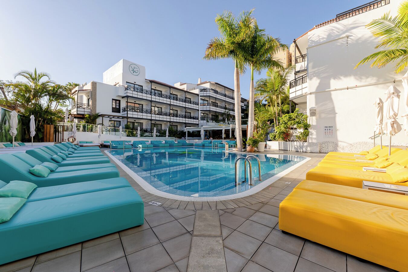 A swimming pool with lounge chairs, a building with balconies, and palm trees.