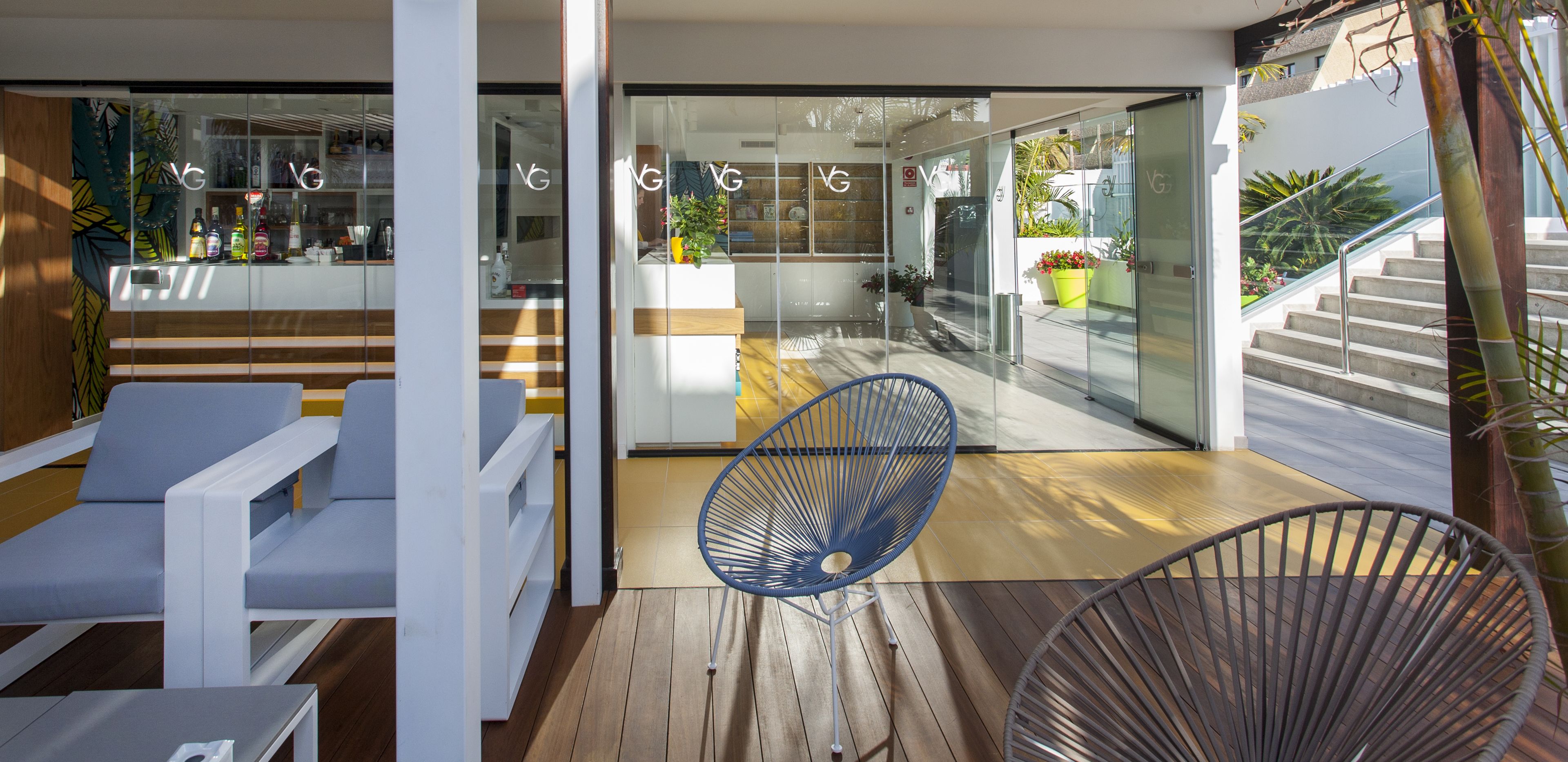 Ein modernes Interieur mit einer Glastür, die in einen Küchenbereich führt, eine Holzterrasse mit Sitzgelegenheiten und eine Treppe mit Glasgeländer.
