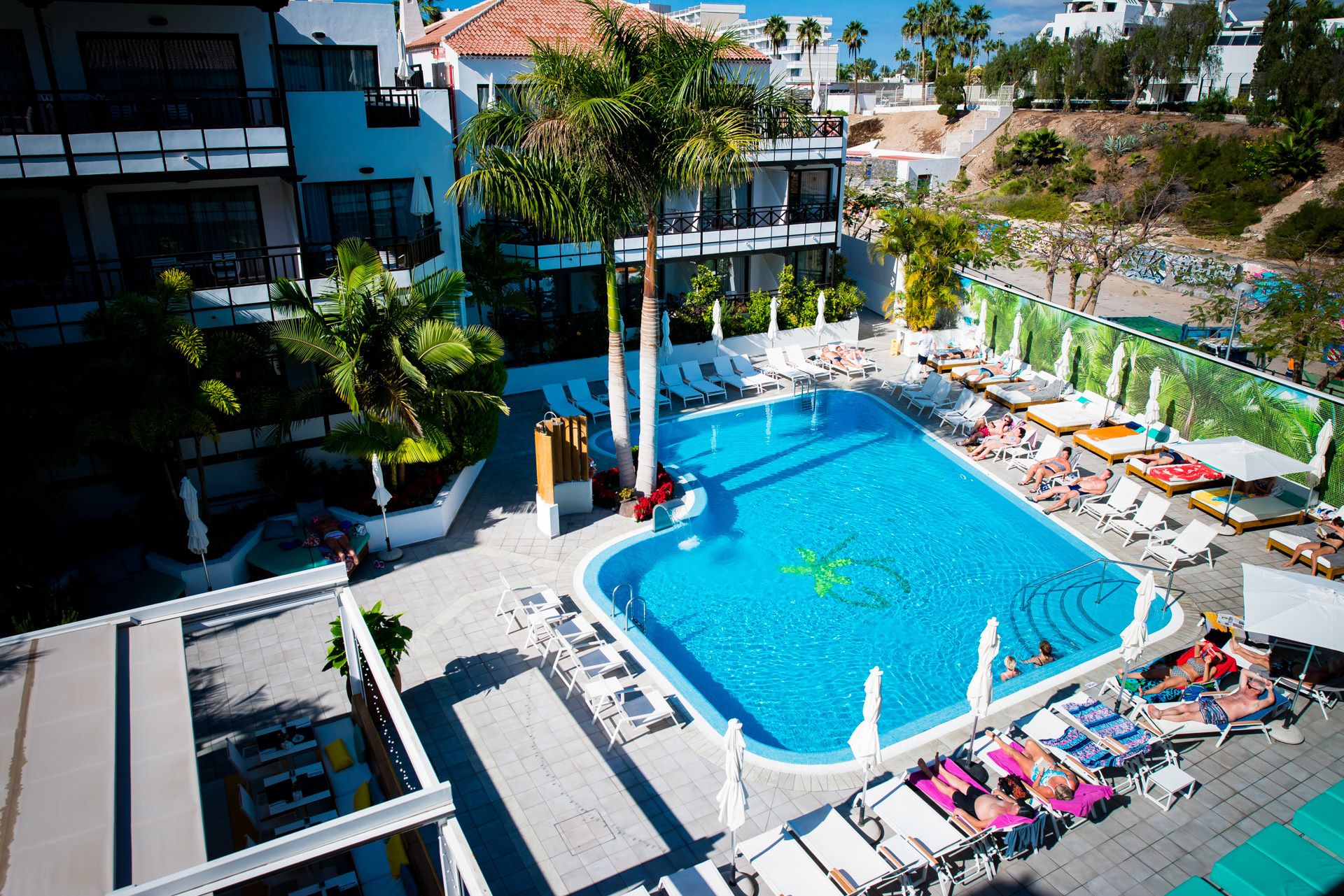 Una piscina en la azotea con tumbonas y sombrillas, rodeada de palmeras y edificios modernos.