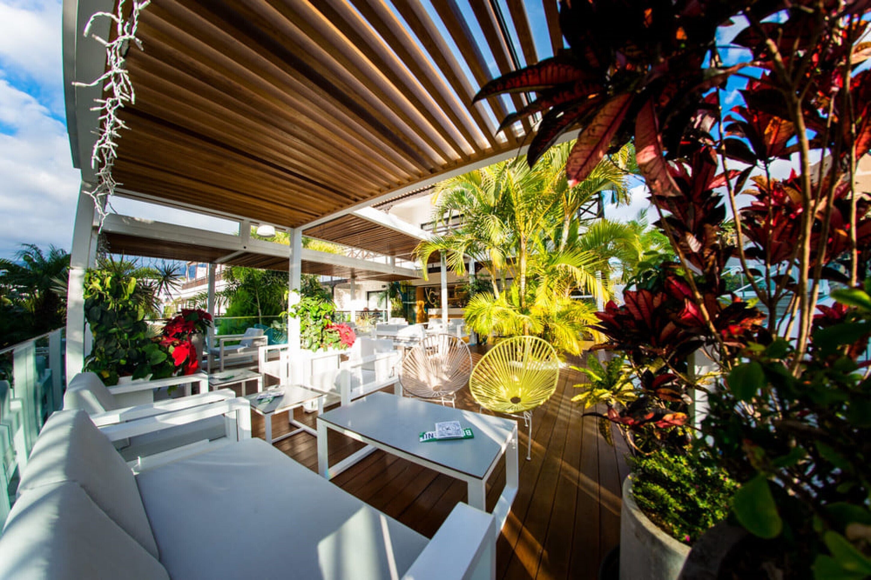 Un patio al aire libre con mobiliario blanco, incluyendo sofás y sillones, bajo una pérgola de madera.