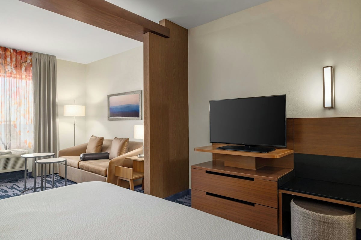 A hotel room with a large bed, a TV on a wooden stand, a desk with drawers, a chair, a lamp, and a view of the outdoors through the window.