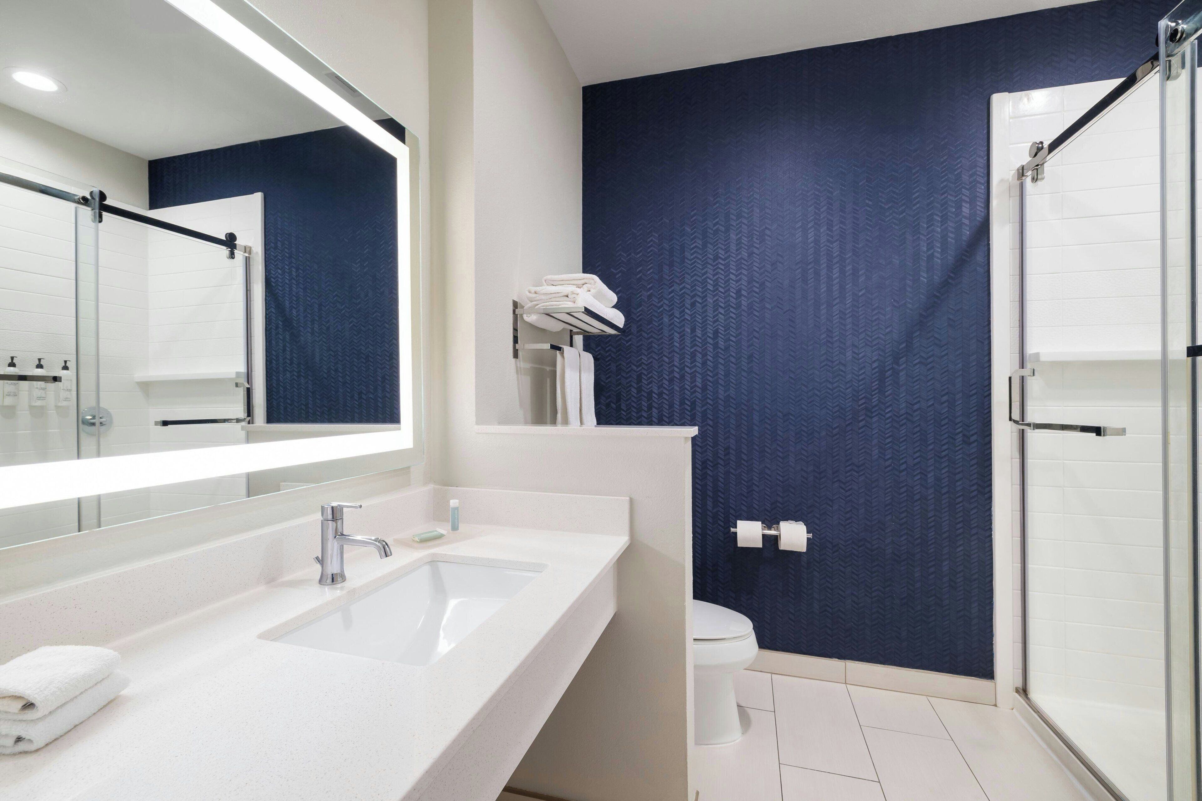 Un baño moderno con un lavamanos blanco, un espejo grande, una ducha y una pared azul oscuro con textura.