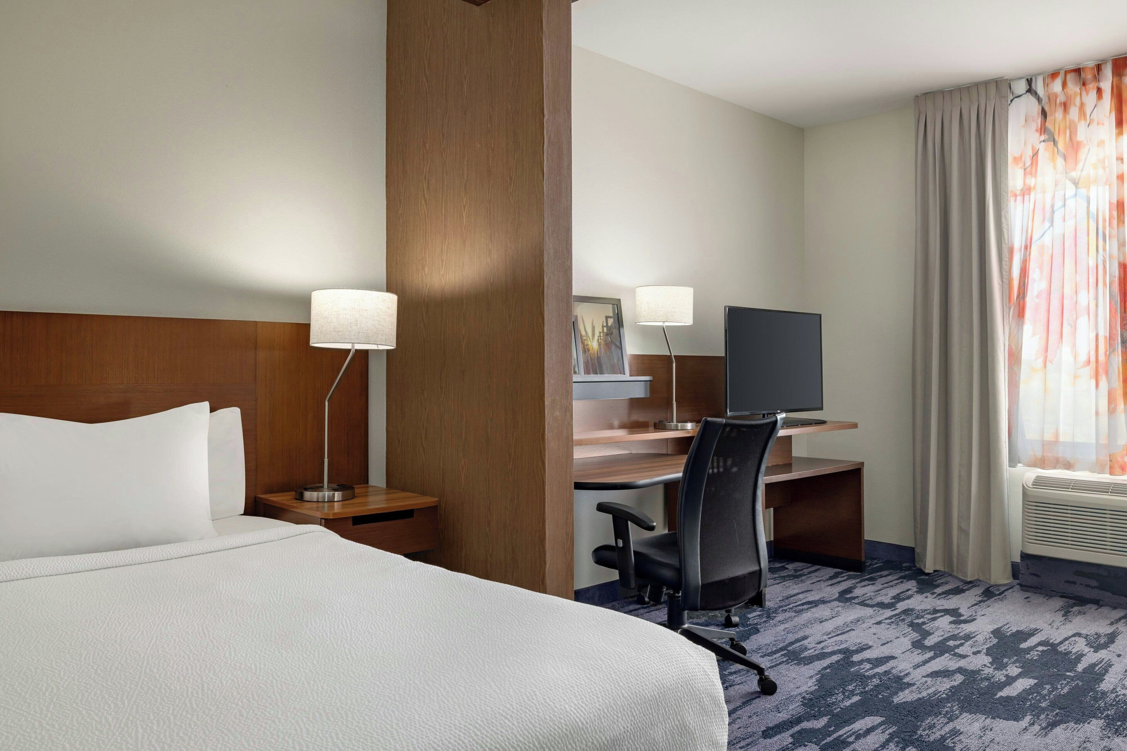 A hotel room with a bed, a desk, a chair, a TV, and a window with curtains.