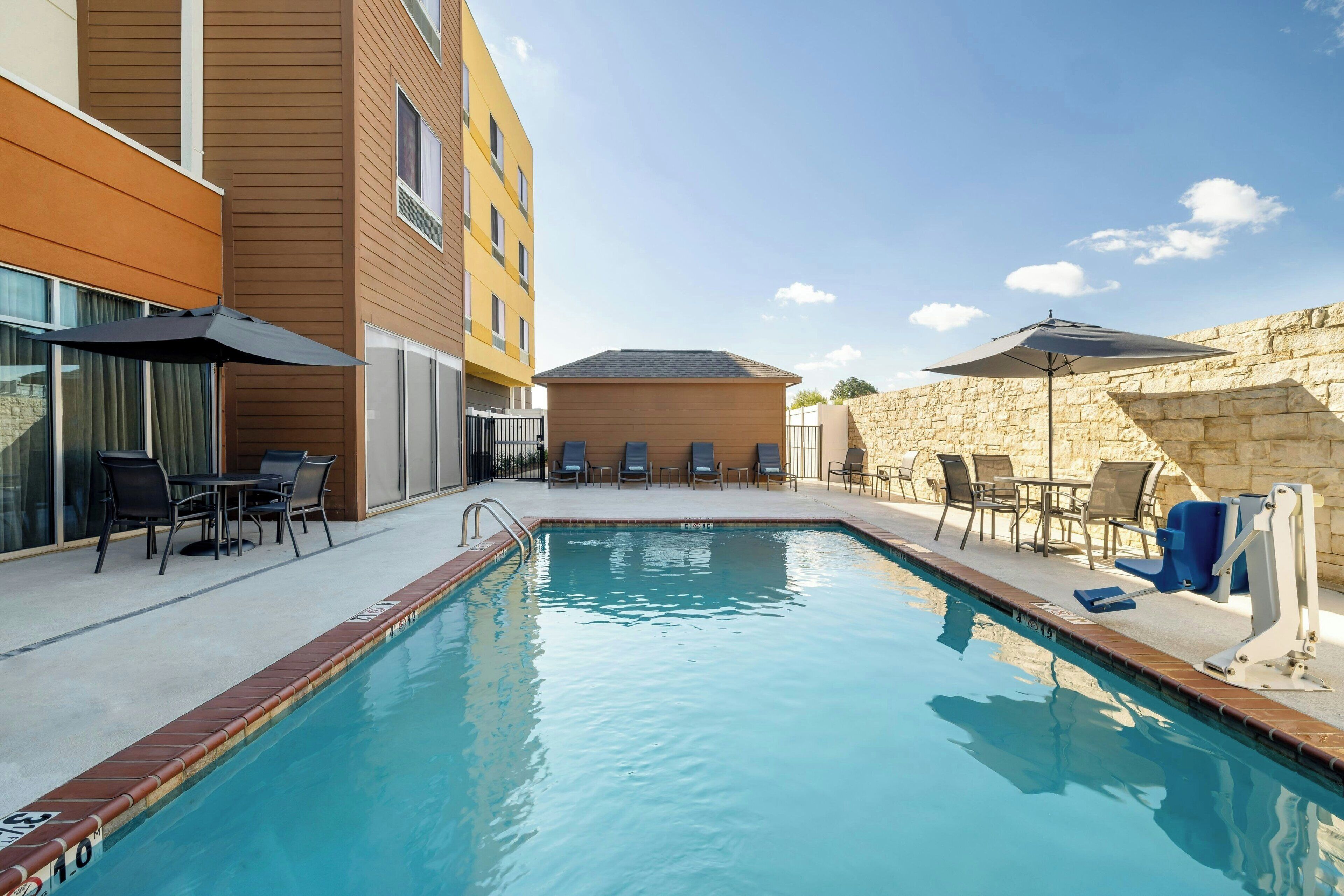 A swimming pool with lounge chairs and umbrellas, surrounded by a stone wall and a building with a flat roof.
