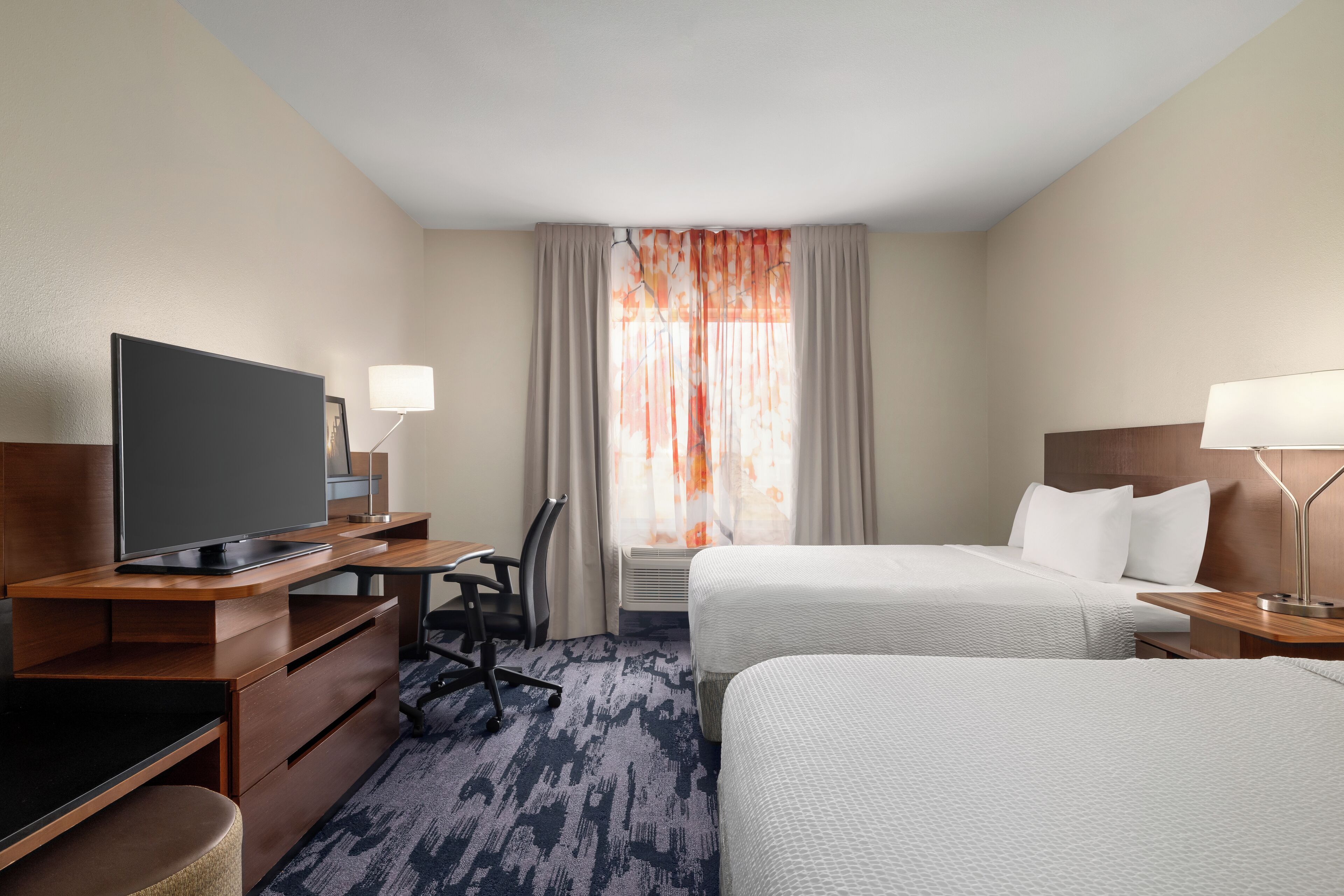 A hotel room with two beds, a desk with a television, a chair, and a lamp.