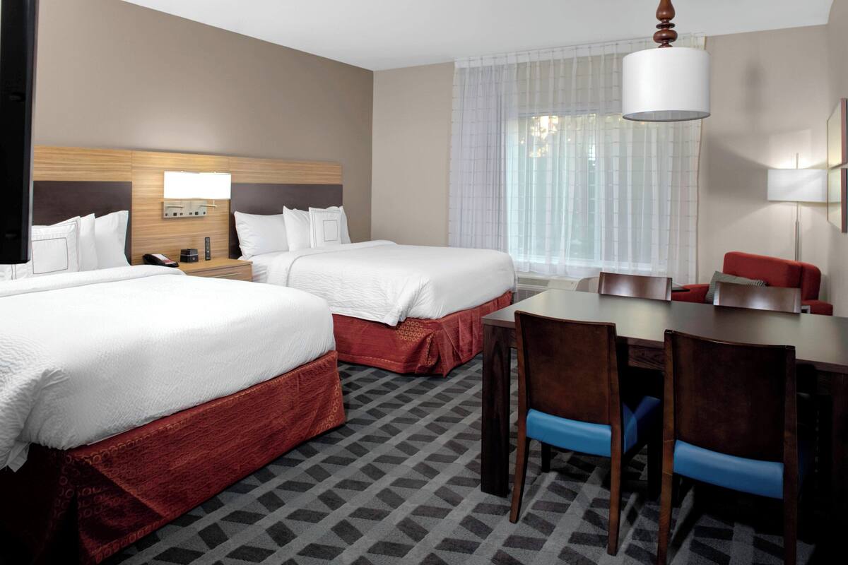 A hotel room with two beds, a dining table, chairs, a TV, a lamp, and a window with curtains.