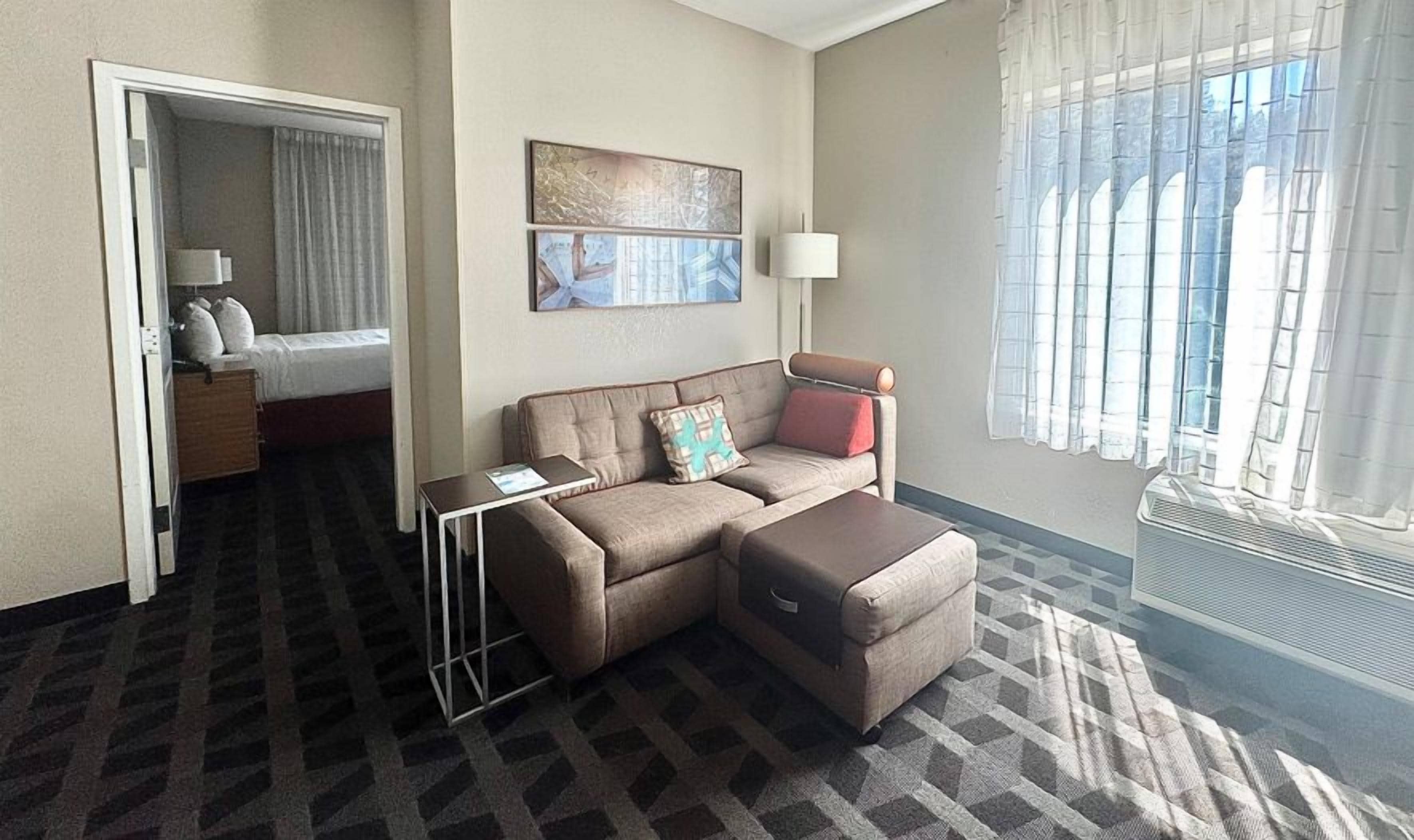 A hotel room with a sofa, ottoman, a small table, a bed, and a window with curtains.