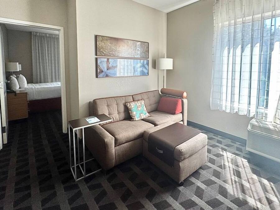A hotel room with a sofa, ottoman, a small table, a bed, and a window with curtains.
