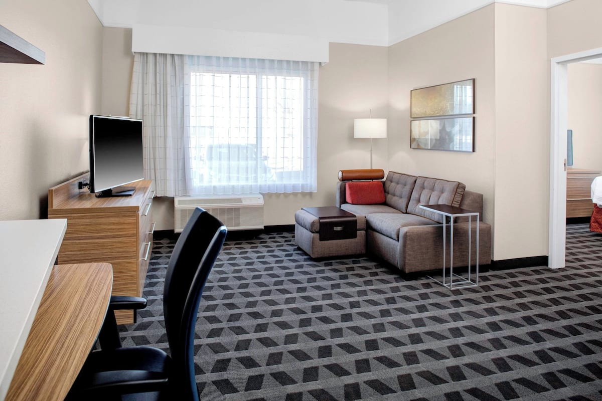 A hotel room with a flat-screen TV, a sofa, a small table, a lamp, and a window with sheer curtains.