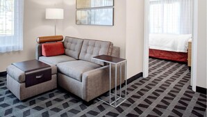 A modern hotel room with a sectional sofa, a small side table, a lamp, and a bed with a red blanket. - TownePlace Suites by Marriott Albany (Albany)