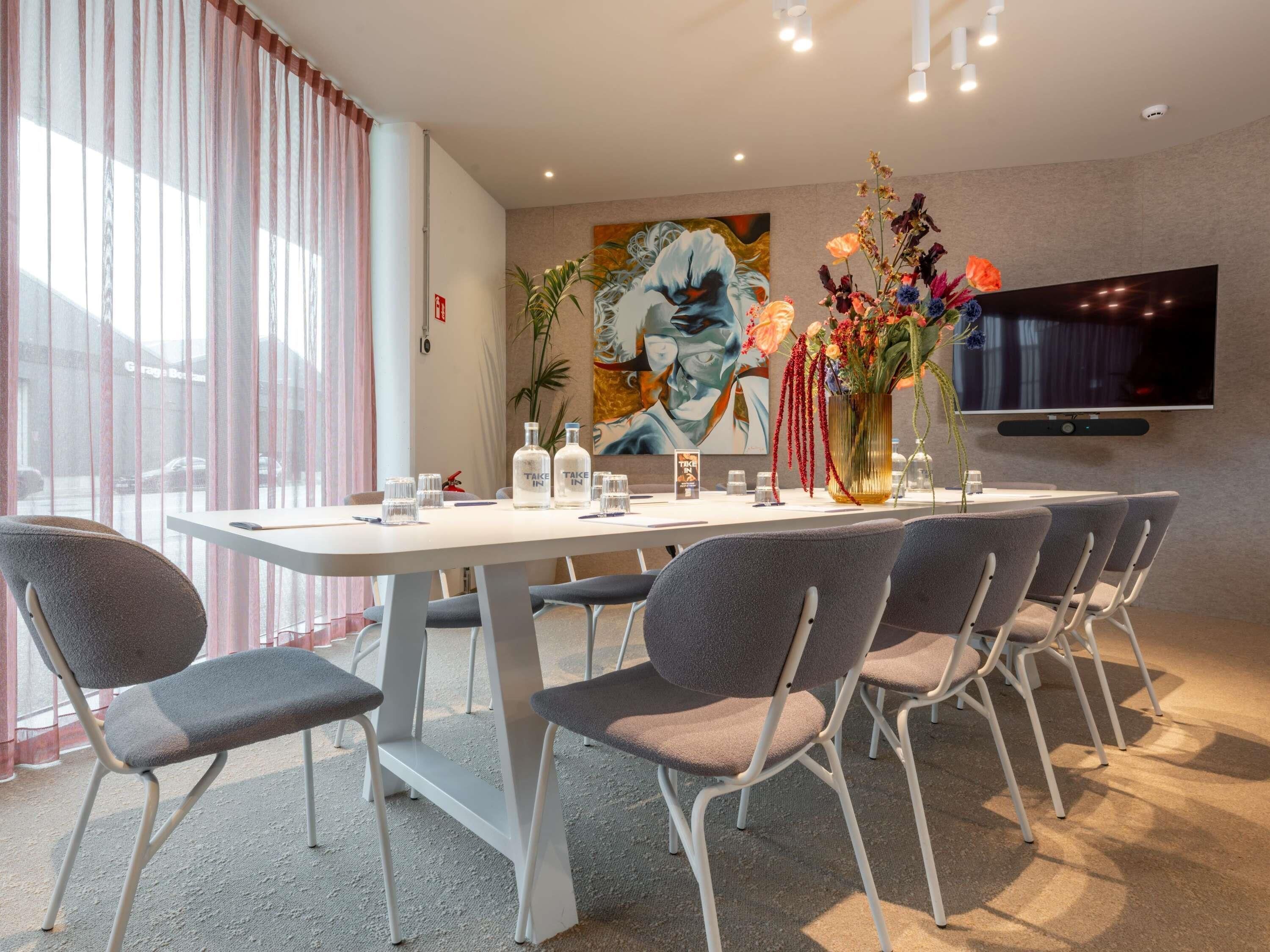 A modern hotel room with a dining area featuring a white table, grey chairs, a flat-screen TV mounted on the wall, and a large floral centerpiece.