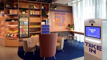 Ein modernes Interieur mit einem Automaten mit der Aufschrift "TAKE IN", Regalen voller Snacks und Getränke sowie einer Sitzecke mit Stühlen und einem Tisch.
