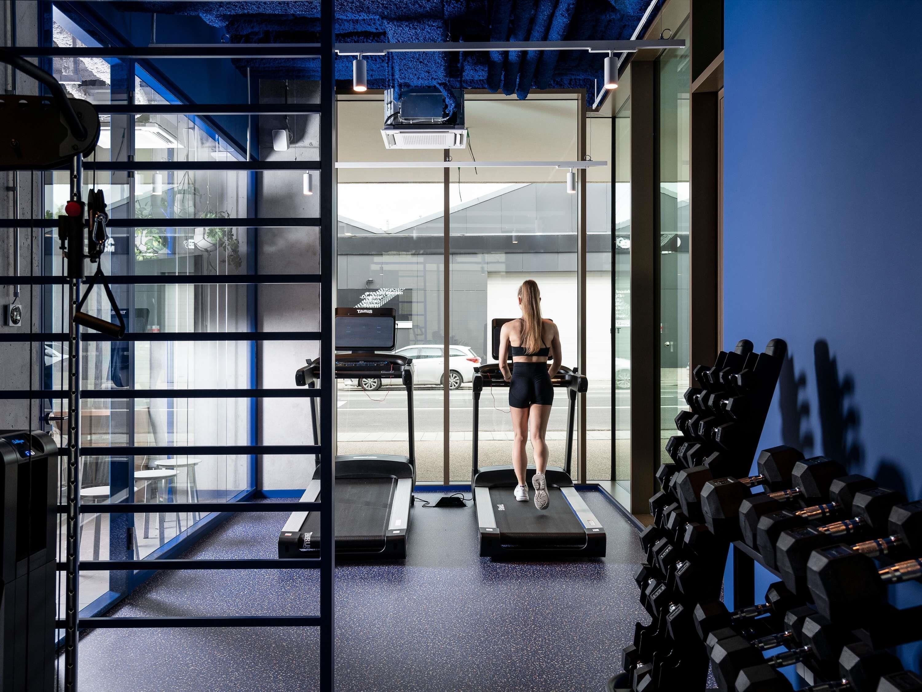 Une salle de sport équipée de tapis de course, d’haltères et dotée d’une grande fenêtre donnant sur un parking.