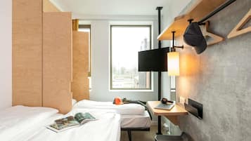 A hotel room with two beds, a desk with a laptop, a chair, a TV mounted on the wall, and a window with a view of buildings.