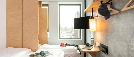 A hotel room with two beds, a desk with a laptop, a chair, a TV mounted on the wall, and a window with a view of buildings.