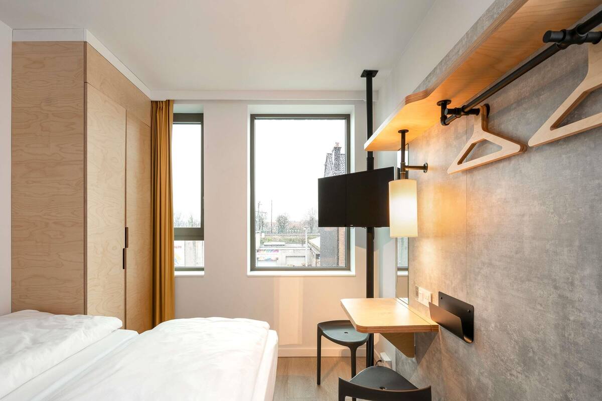 Habitación de hotel con cama, escritorio, silla, televisión y ventana con vista a edificios.